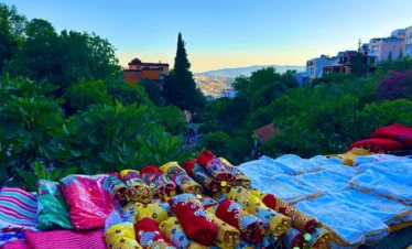 Colorful sequined fabrics at a stall by Ras El Maa in Chefchaouen, overlooking a lush ravine and hillside houses beneath a clear blue sky.