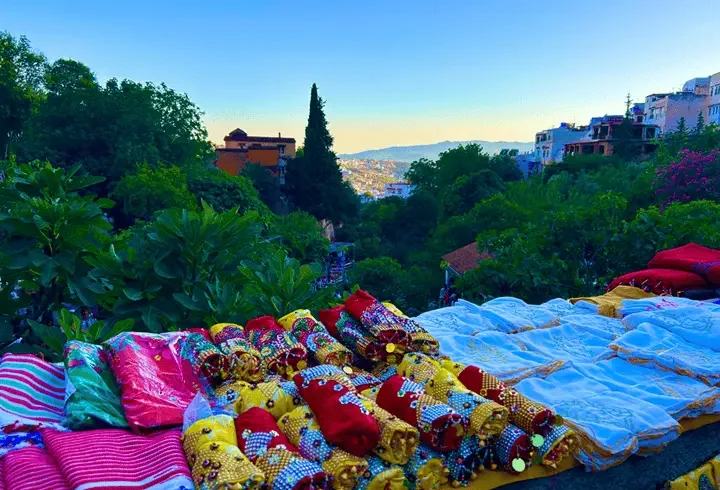 Colorful sequined fabrics at a stall by Ras El Maa in Chefchaouen, overlooking a lush ravine and hillside houses beneath a clear blue sky.