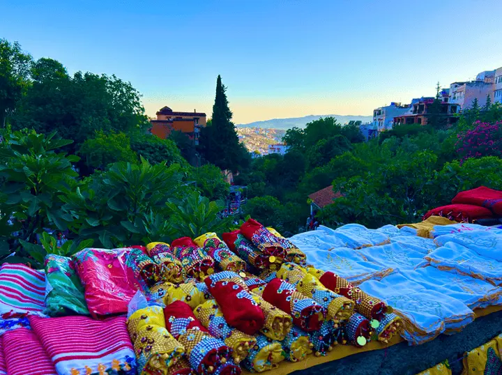Colorful sequined fabrics at a stall by Ras El Maa in Chefchaouen, overlooking a lush ravine and hillside houses beneath a clear blue sky.