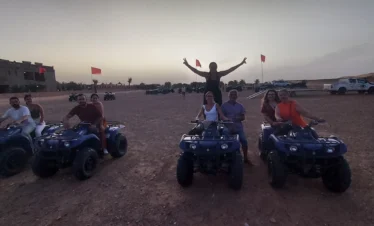 At dusk in the Zagora desert, a group poses on blue quad bikes while a person stands with arms raised behind them, with red flags, tents, and 4x4s in the background.