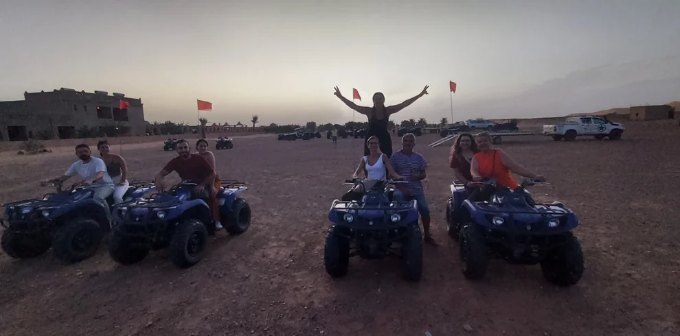 At dusk in the Zagora desert, a group poses on blue quad bikes while a person stands with arms raised behind them, with red flags, tents, and 4x4s in the background.