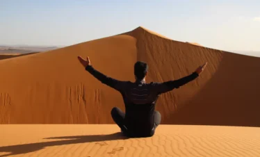 Back view of a traveler sitting cross-legged on the golden dunes of Erg Chebbi near Merzouga, Morocco, arms outstretched toward a high ridge under a clear sky, footprints behind.