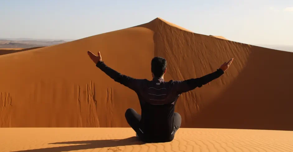 Back view of a traveler sitting cross-legged on the golden dunes of Erg Chebbi near Merzouga, Morocco, arms outstretched toward a high ridge under a clear sky, footprints behind.