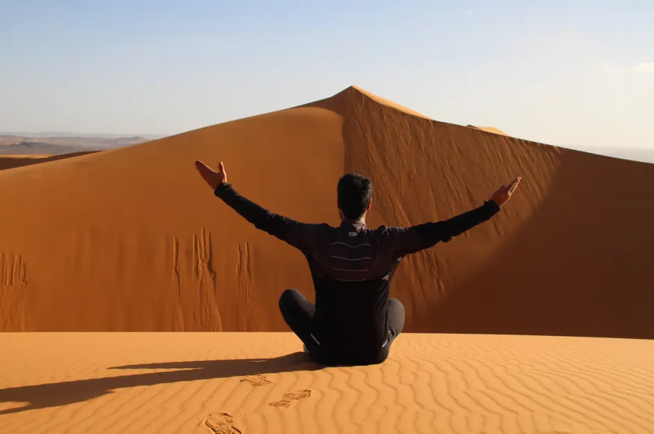 Back view of a traveler sitting cross-legged on the golden dunes of Erg Chebbi near Merzouga, Morocco, arms outstretched toward a high ridge under a clear sky, footprints behind.