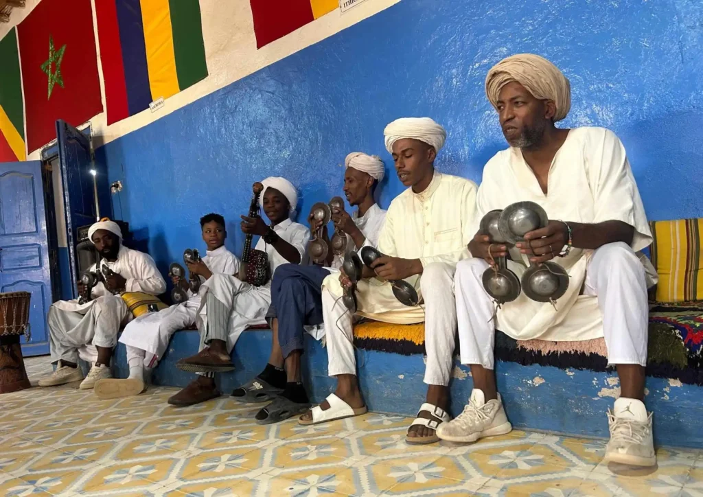 Gnawa musicians in white robes and turbans sit along a blue wall, playing metal qraqeb castanets and a guembri lute in a tiled room decorated with flags.