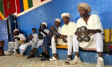 Gnawa musicians in white robes and turbans sit along a blue wall, playing metal qraqeb castanets and a guembri lute in a tiled room decorated with flags.