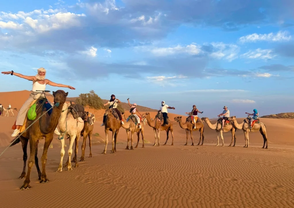 Caravan of tourists riding camels in a line across the golden dunes of Erg Chebbi near Merzouga, arms outstretched under a blue, cloud-streaked sky.