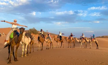Caravan of tourists riding camels in a line across the golden dunes of Erg Chebbi near Merzouga, arms outstretched under a blue, cloud-streaked sky.