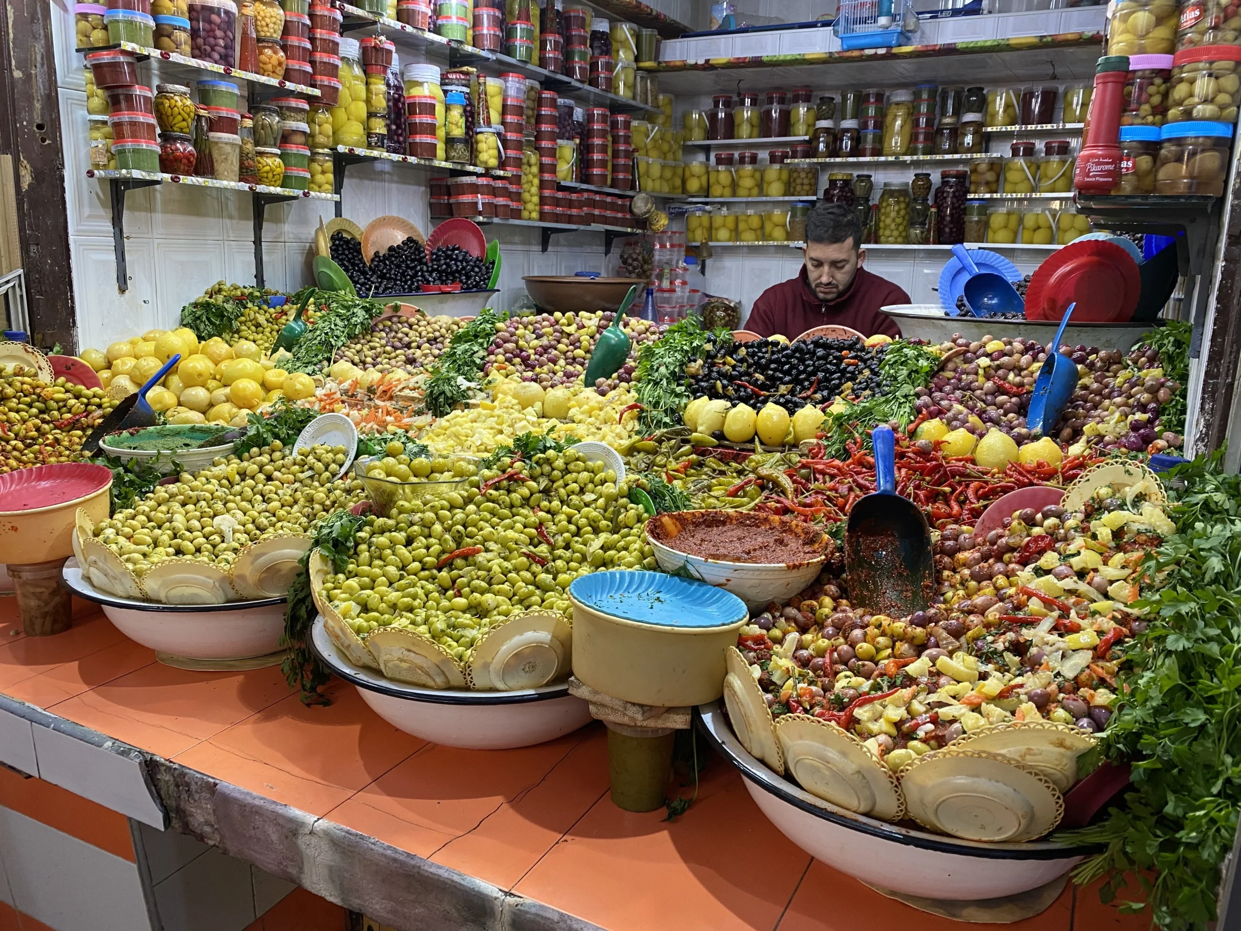 Colorful olive stand in a Moroccan souk with mounds of olives, preserved lemons, red chiles, and fresh herbs.