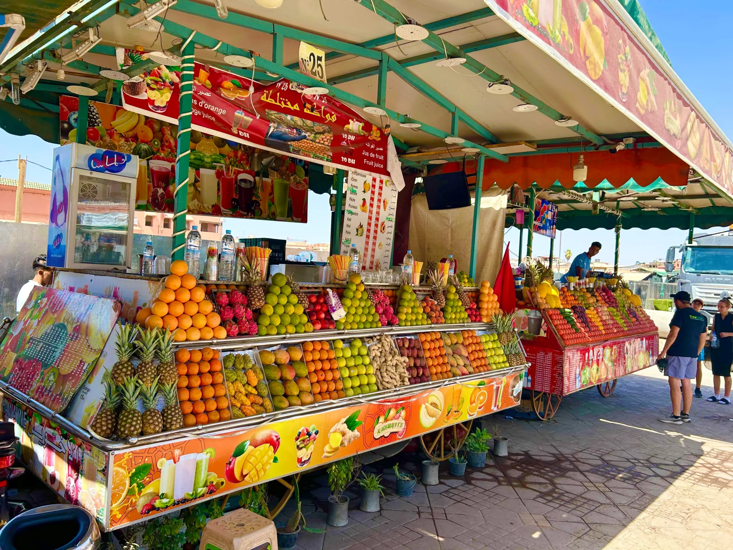 Fruit juice stalls at Jemaa el-Fnaa, Marrakech, stacked pyramids of oranges, mangoes and pineapples under a green awning.