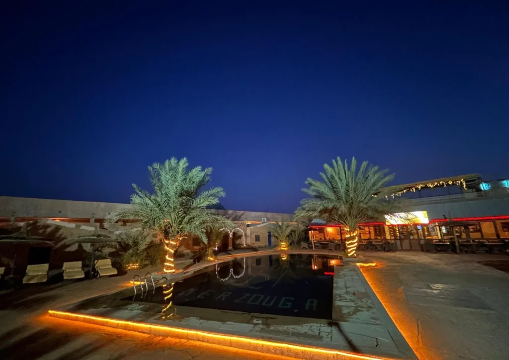 Palm trees wrapped in lights flank a reflective pool in a desert lodge courtyard, warm-lit seating around it under a deep indigo sky.