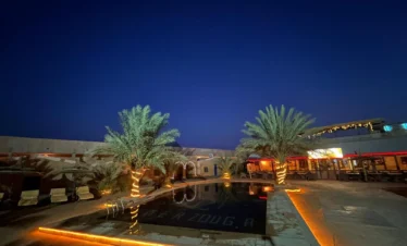 Palm trees wrapped in lights flank a reflective pool in a desert lodge courtyard, warm-lit seating around it under a deep indigo sky.