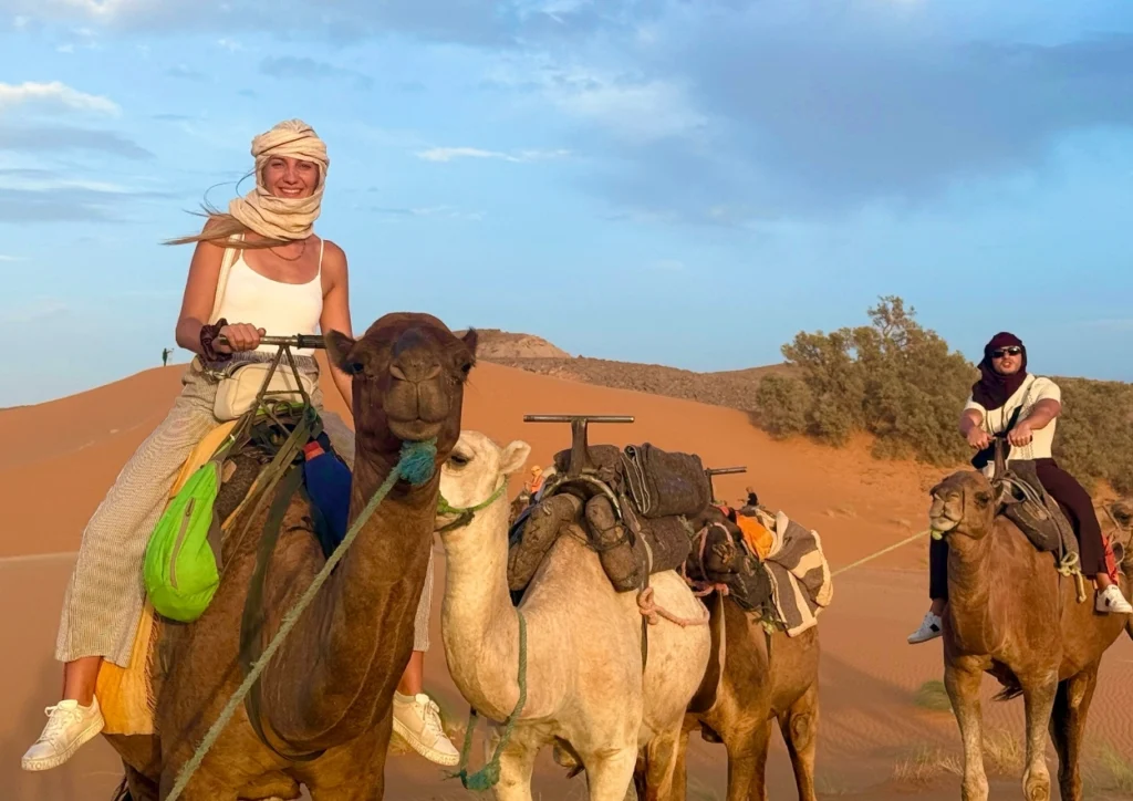 Smiling traveler in a headscarf rides the lead camel as a small caravan crosses orange Sahara dunes, another rider following under a pale blue evening sky.