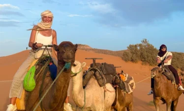 Smiling traveler in a headscarf rides the lead camel as a small caravan crosses orange Sahara dunes, another rider following under a pale blue evening sky.