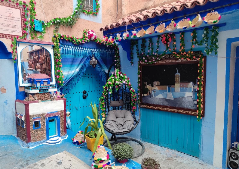 Colorful blue-washed corner in Chefchaouen with decorated arched door, hanging chair, flower garlands and woven baskets.