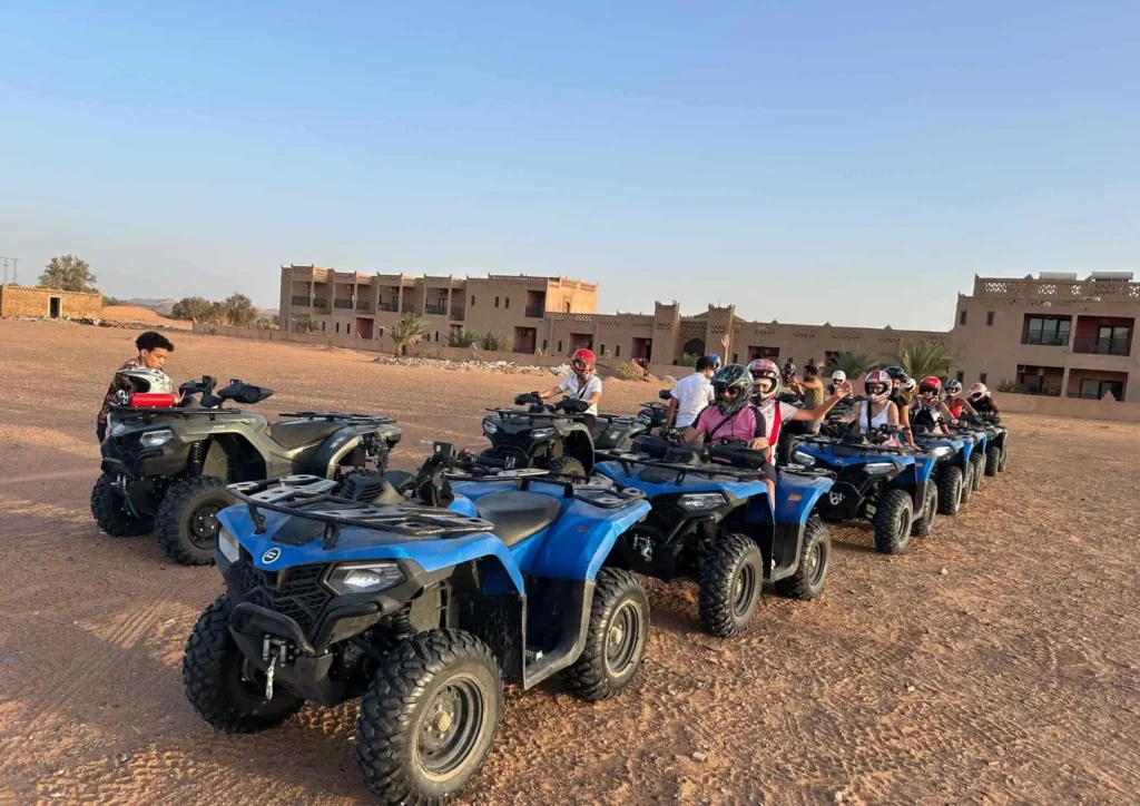 Row of blue ATVs with helmeted riders lined up on a desert plain outside a kasbah-style hotel, ready for a quad tour under a clear sky.
