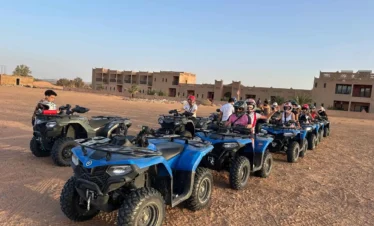Row of blue ATVs with helmeted riders lined up on a desert plain outside a kasbah-style hotel, ready for a quad tour under a clear sky.