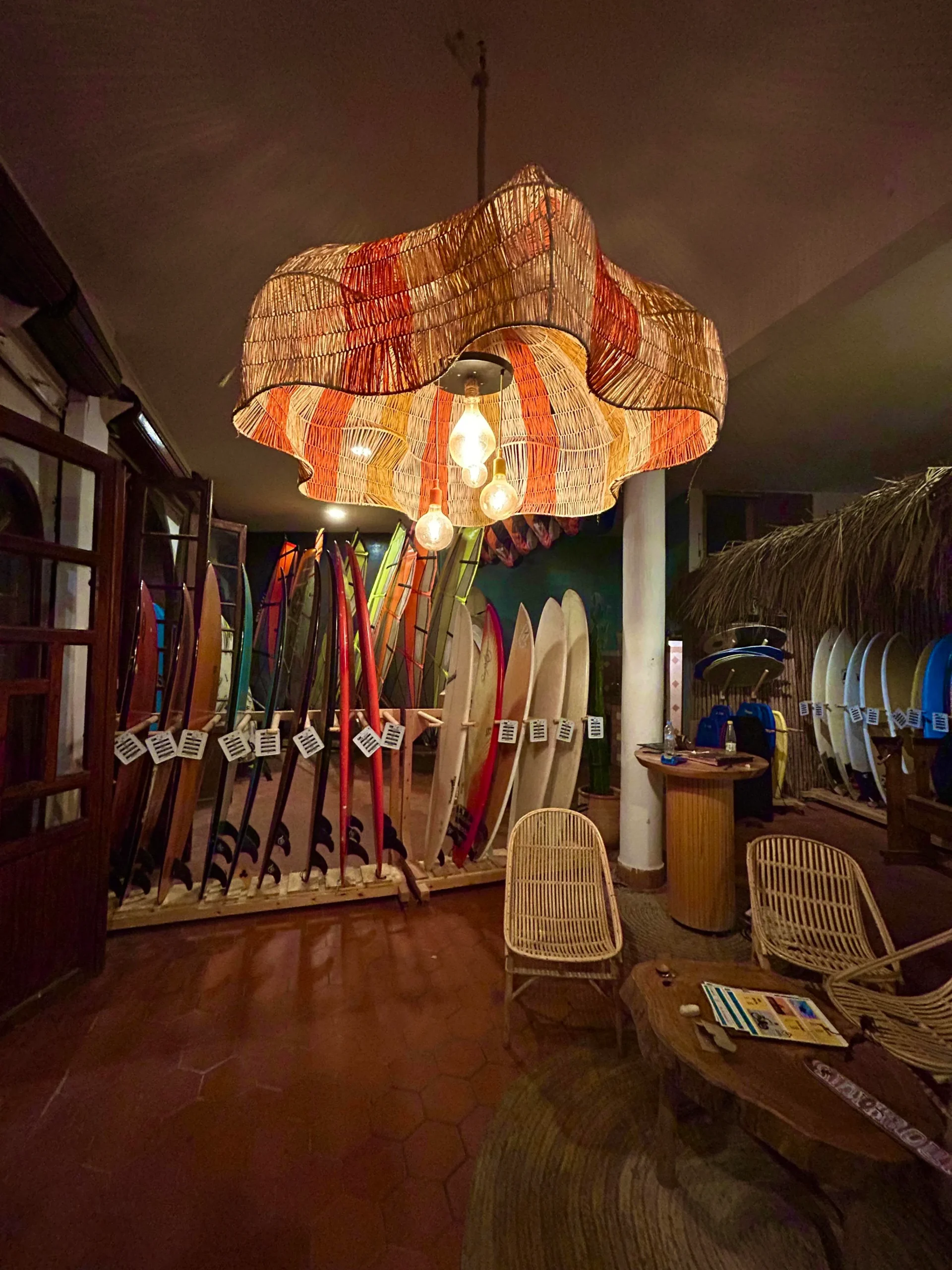 Surf club lounge with rows of colorful surfboards, a woven pendant lamp, and rattan chairs in warm evening light.