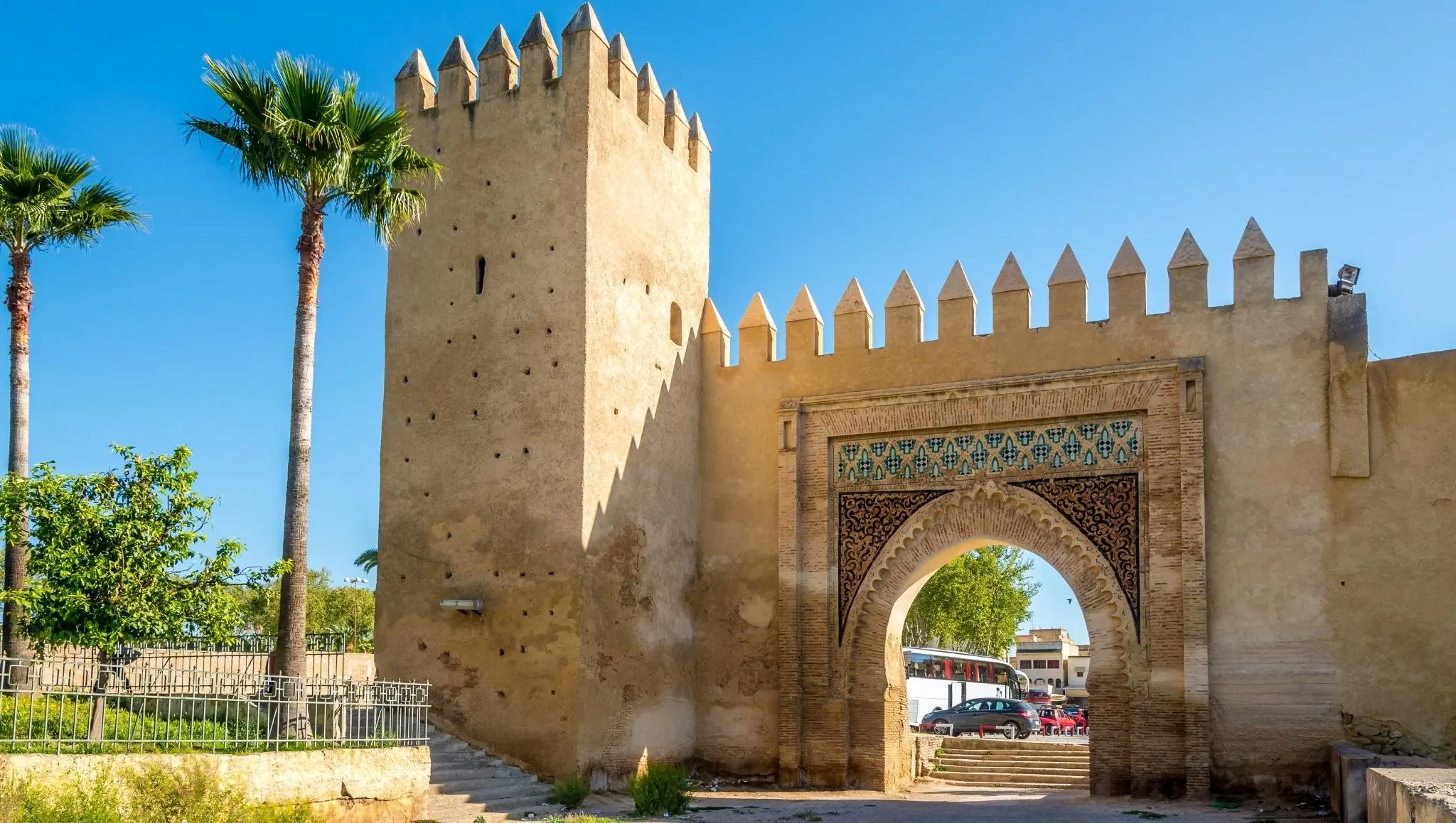 Sunlit view of a crenellated city gate with decorative tilework and palm trees—typical medina entrance included in a 2 days Fes itinerary.