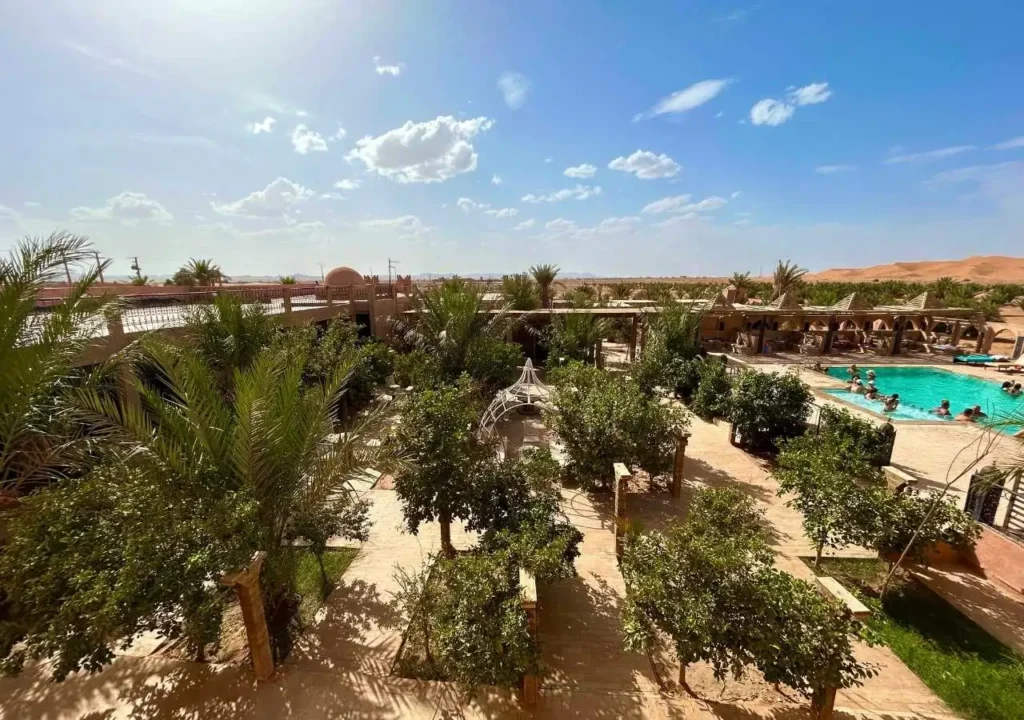 Aerial view of palm-filled courtyard with swimming pool and Erg Chebbi dunes in the distance.