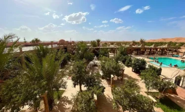 Aerial view of palm-filled courtyard with swimming pool and Erg Chebbi dunes in the distance.
