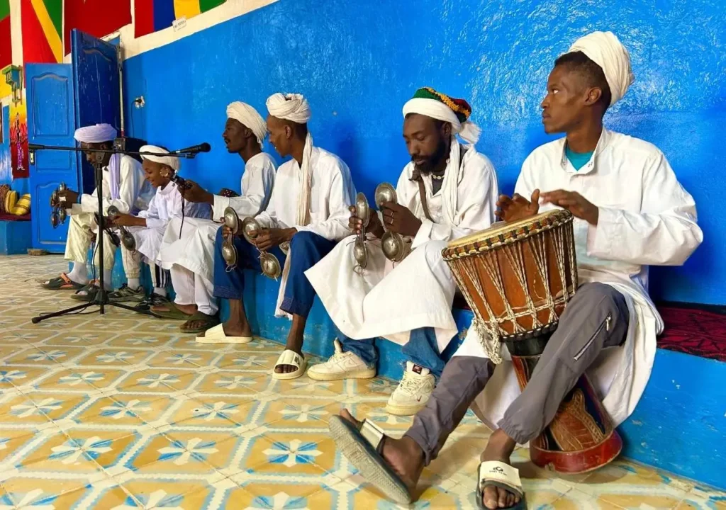 Gnawa band in white robes playing krakebs and drum inside a vivid blue room in Khamlia village.