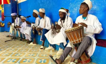 Gnawa band in white robes playing krakebs and drum inside a vivid blue room in Khamlia village.