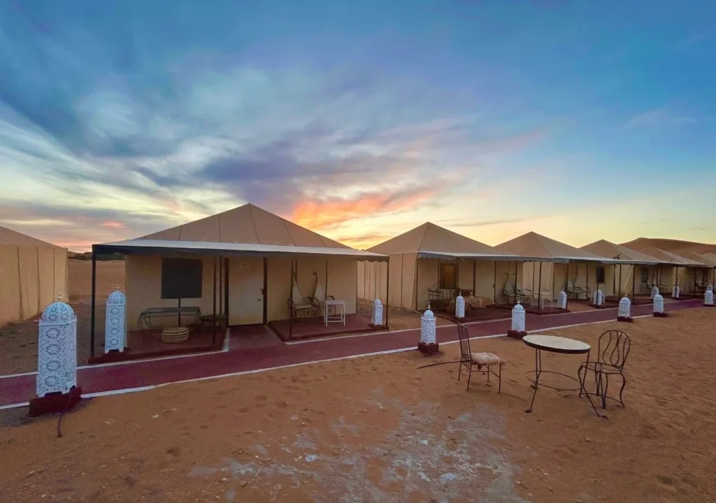 Row of private desert tents with lanterns glowing at dusk on the sands of Merzouga.