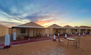 Row of private desert tents with lanterns glowing at dusk on the sands of Merzouga.