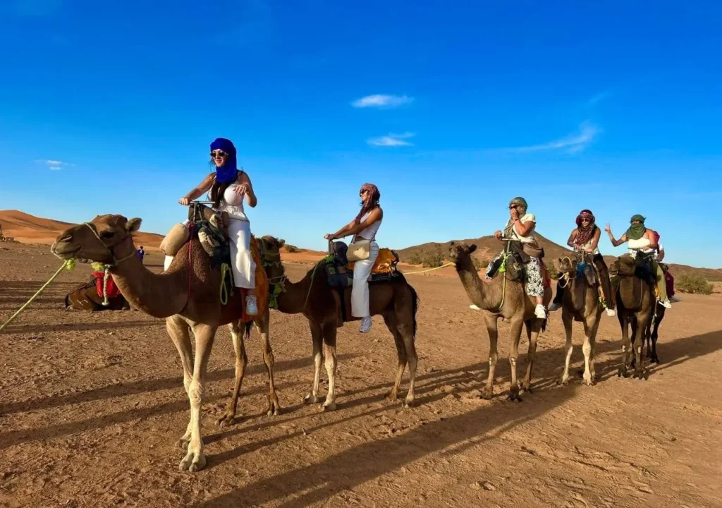 Group of travelers riding camels across golden Merzouga dunes under a bright blue sky.