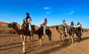 Group of travelers riding camels across golden Merzouga dunes under a bright blue sky.