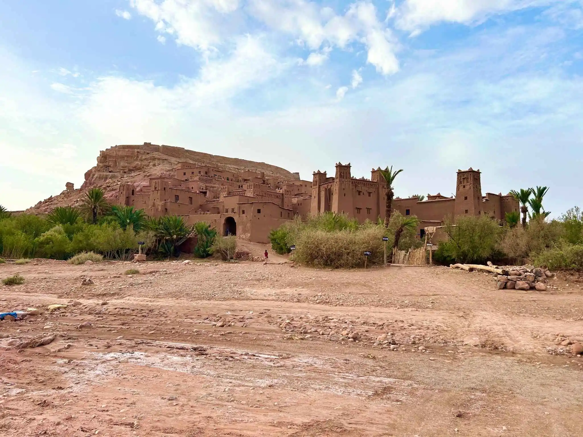 Wide view of the UNESCO-listed ksar of Ait Benhaddou—mud-brick towers and houses climbing a hillside, with palms and a rocky desert foreground beneath scattered clouds.