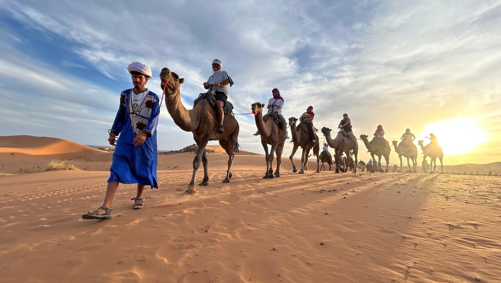 Berber guide leading a line of tourists on camels across the golden dunes of Erg Chebbi at sunset during a Fes to Marrakech camel ride.