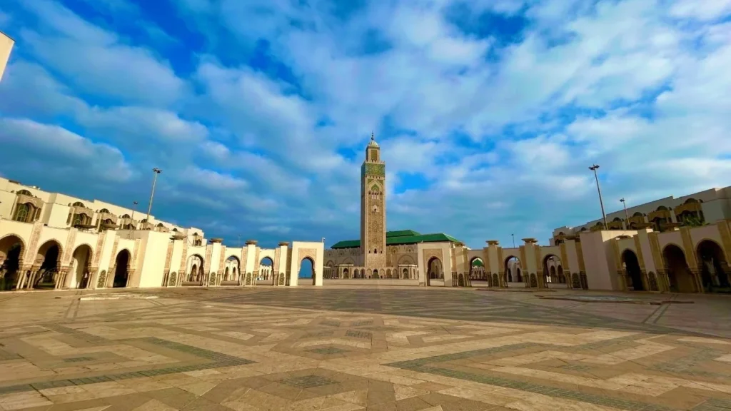 Panoramic view of Hassan II Mosque with its minaret and arcaded esplanade under a blue, cloud-dotted sky.
