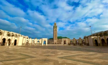 Panoramic view of Hassan II Mosque with its minaret and arcaded esplanade under a blue, cloud-dotted sky.