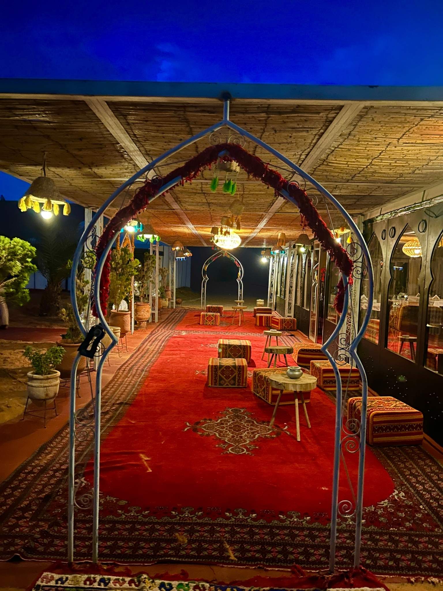 Lantern-lit Berber desert camp with red carpets and low tables glowing at blue hour in Merzouga, Morocco—on a Fes to Marrakech desert tour.