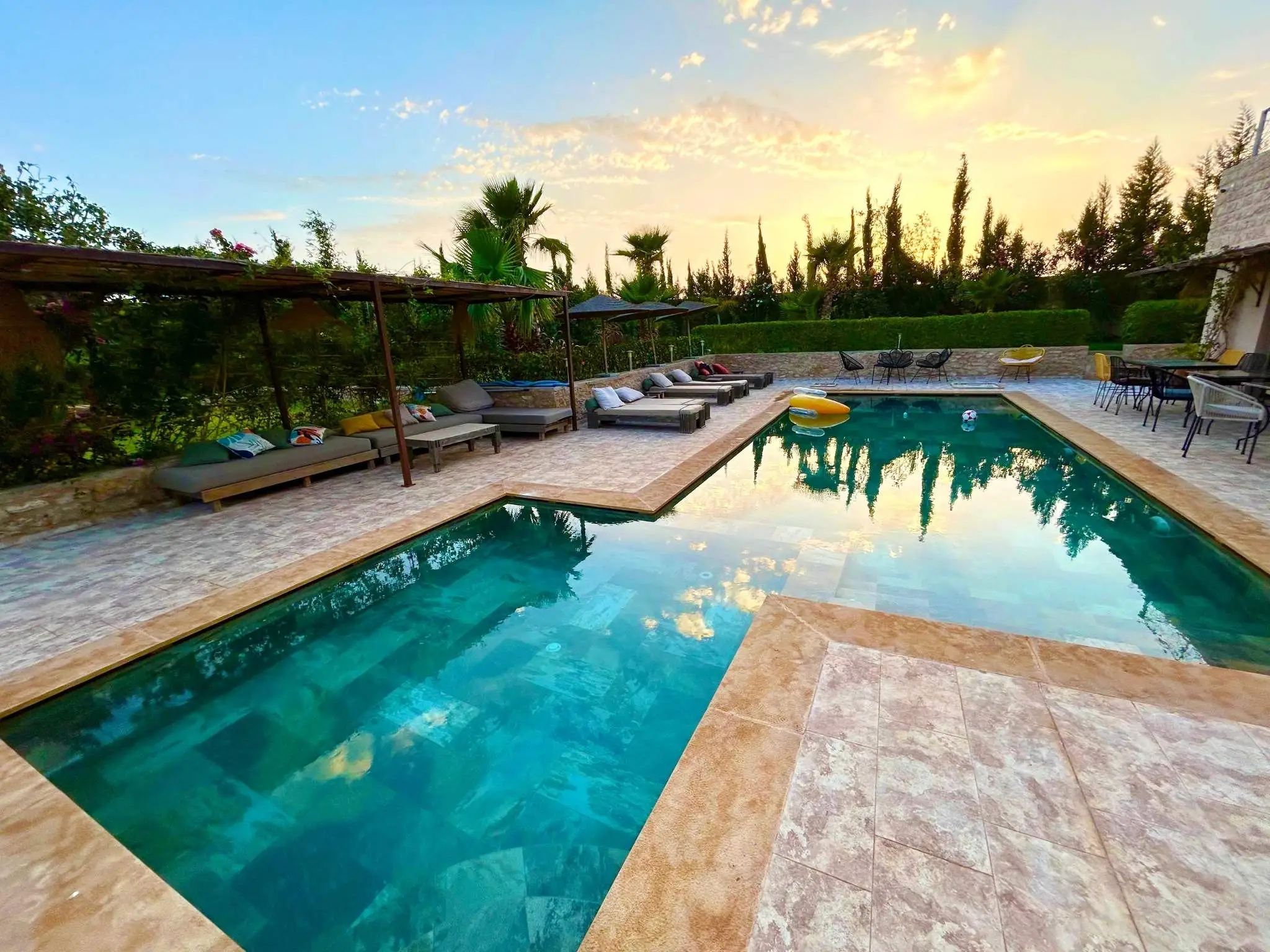 Tranquil riad pool in Essaouira, Morocco at sunset with loungers and palm trees reflecting on turquoise water.