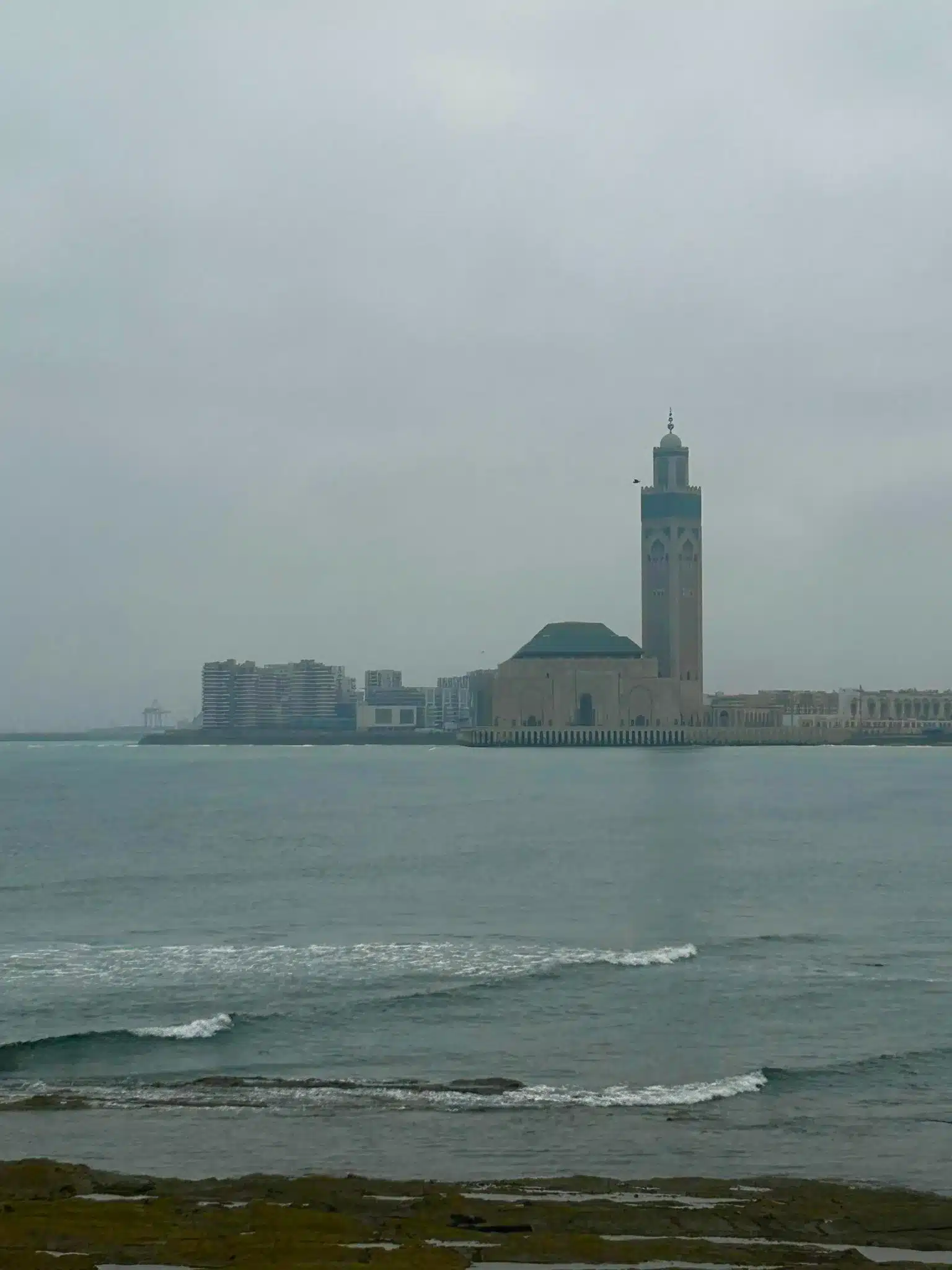 Distant coastal view of the Hassan II Mosque rising above the Atlantic in Casablanca on a hazy day, highlighting the city’s waterfront.