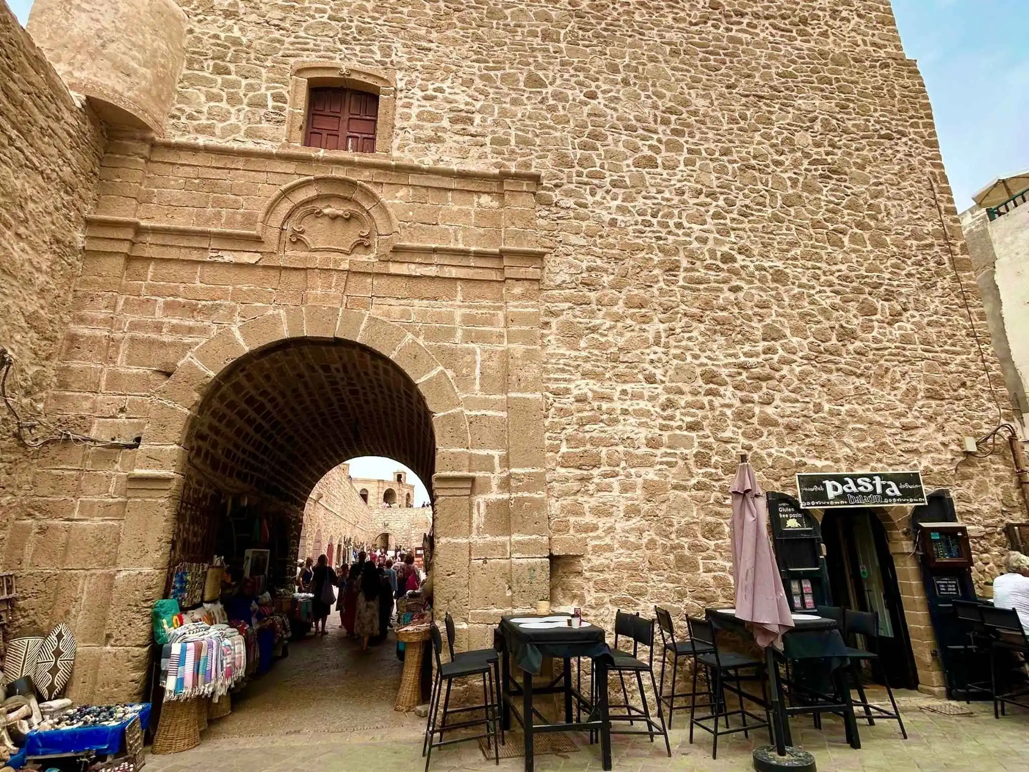 Massive stone gateway and arches leading into Essaouira medina, Morocco, with cafés and market stalls nearby.