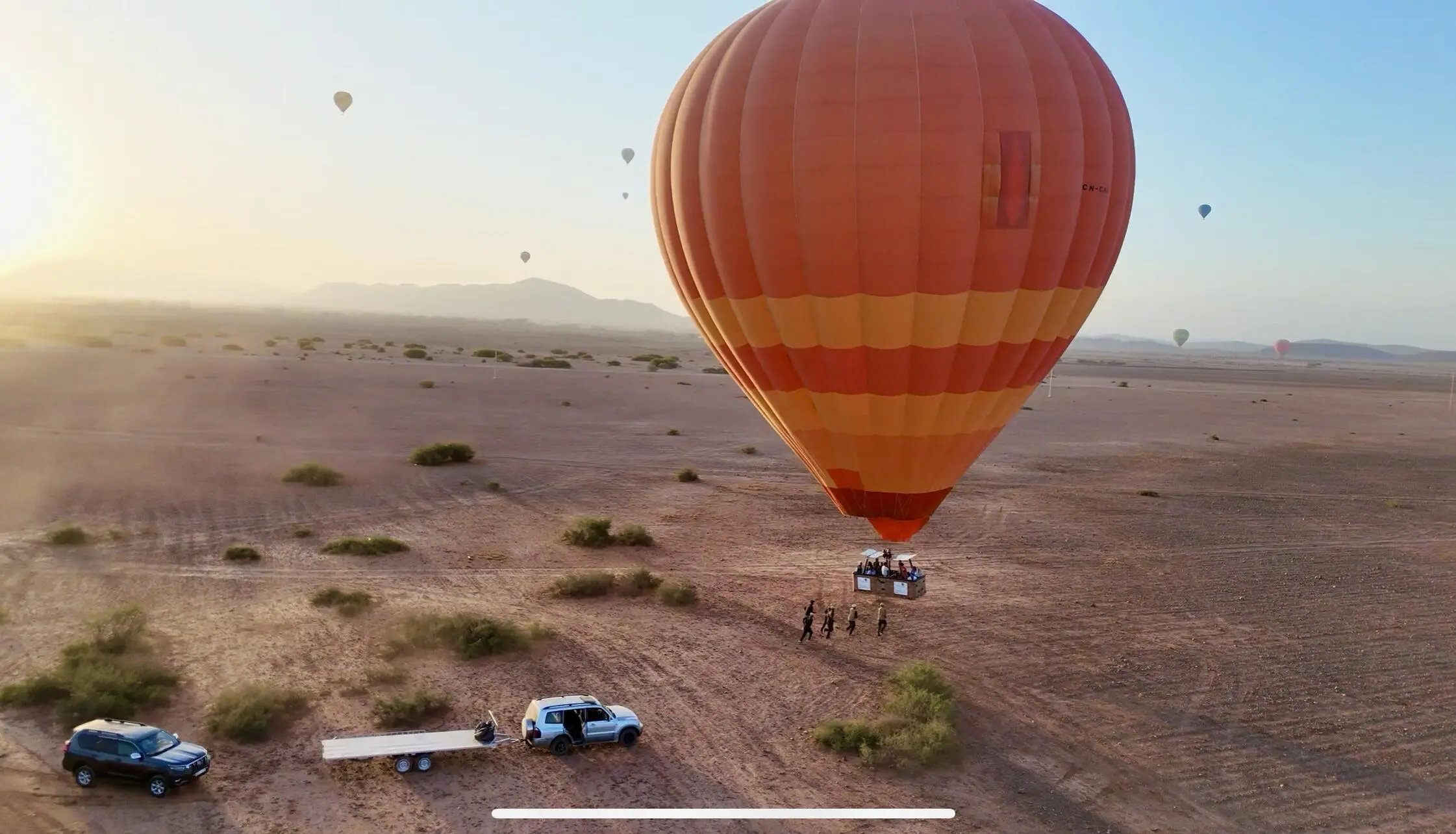 Orange hot air balloon touching down on the Marrakech desert as crew and 4x4 support vehicles assist with the landing.