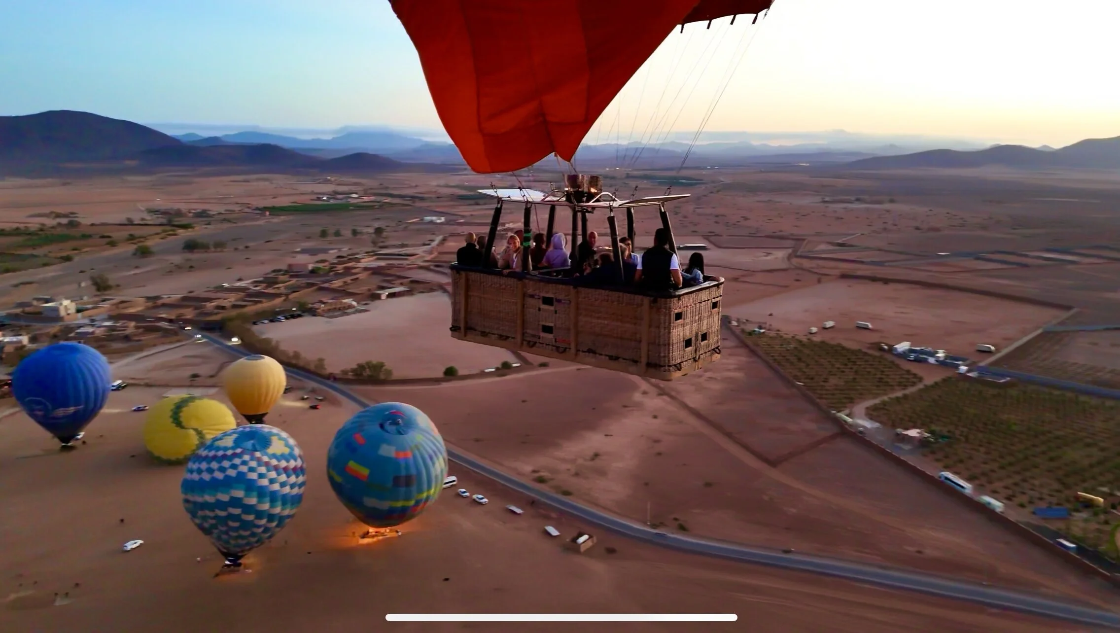 Close-up of a wicker basket full of passengers floating above the Marrakech desert at sunrise, with colorful balloons inflating on the ground below.