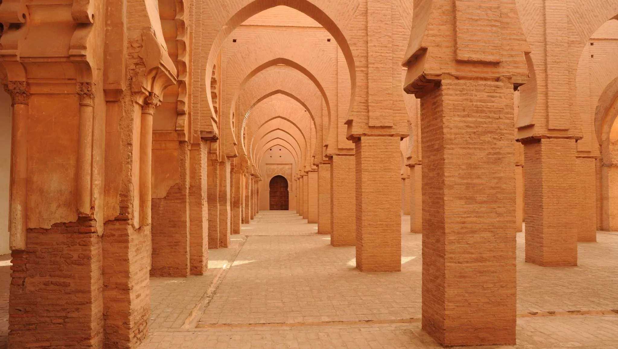 Long corridor of warm brick arches and pillars in Marrakech showcasing classic Almohad architecture and repeating geometric lines.