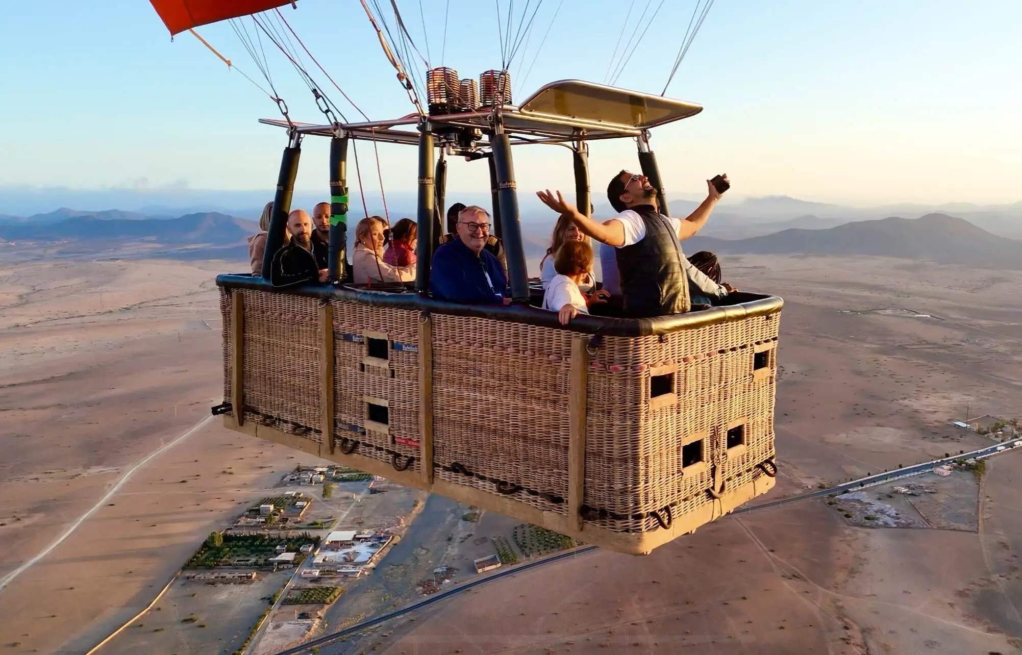 People smiling in a wicker balloon basket during a sunrise flight above the Marrakech desert, with hills and villages below.