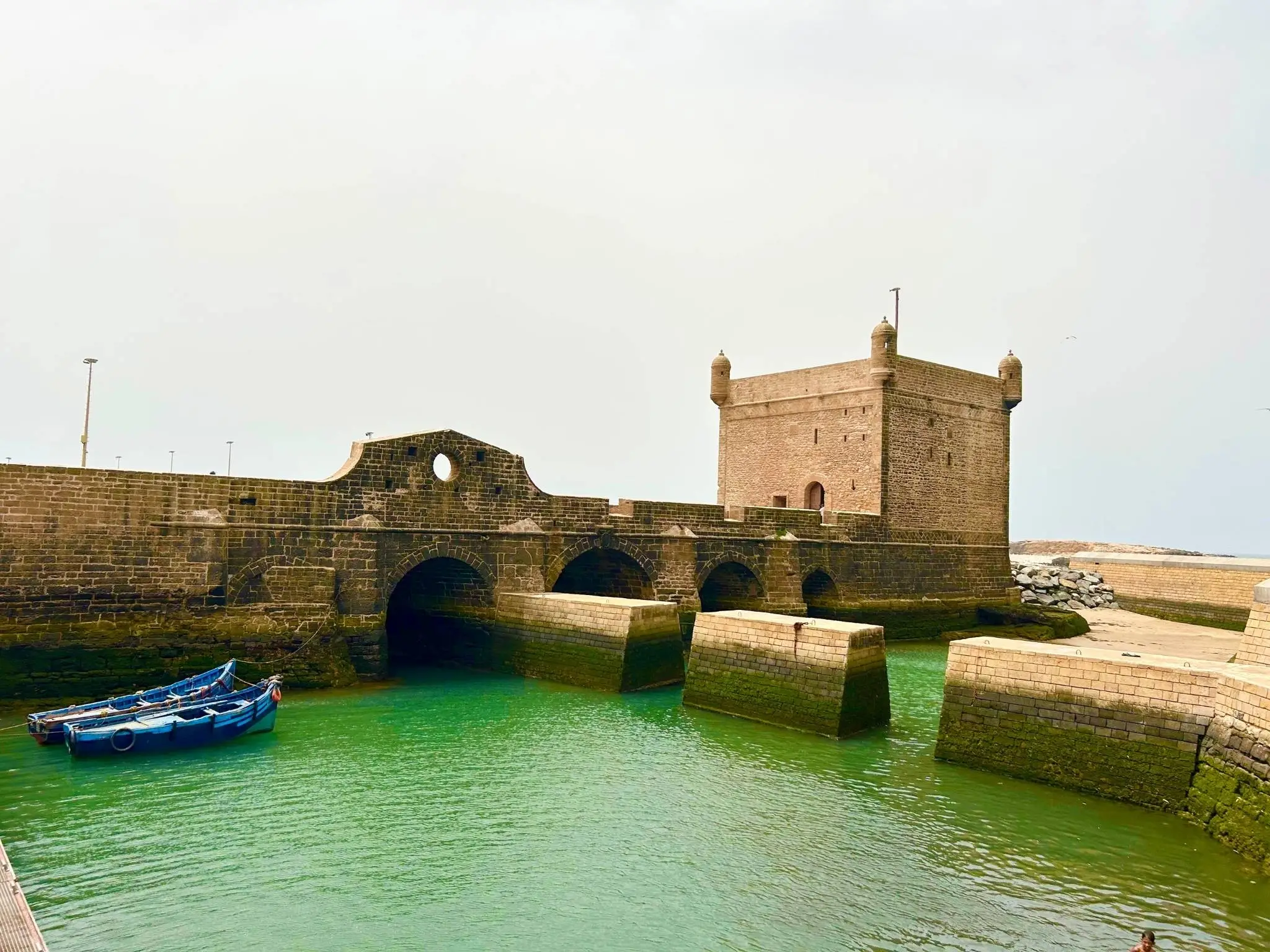 Stone ramparts and watchtower of the Skala de la Ville in Essaouira, Morocco with blue fishing boat on emerald water.