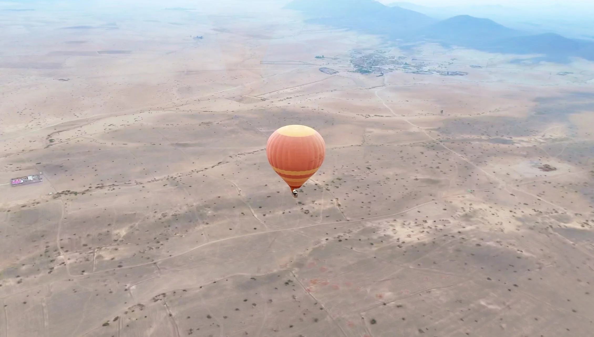 Orange hot air balloon drifting over the Marrakech desert plain with scattered shrubs and small villages; mountains faint in the distance.