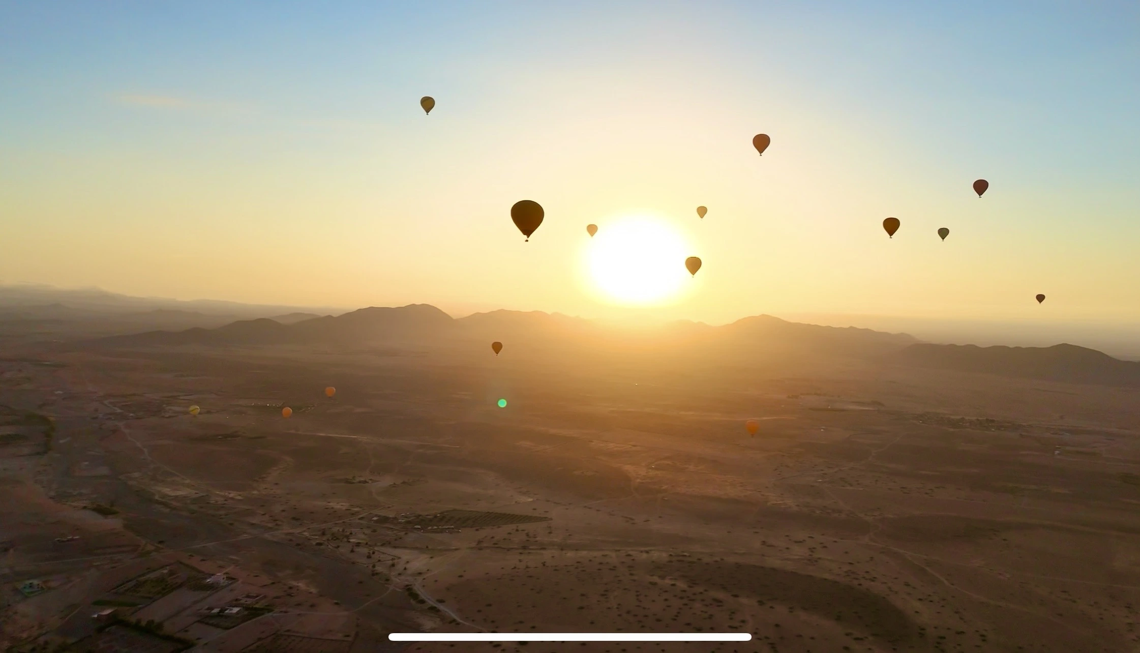 Multiple hot air balloons silhouetted against sunrise above the Marrakech desert, the Atlas Mountains visible on the horizon.