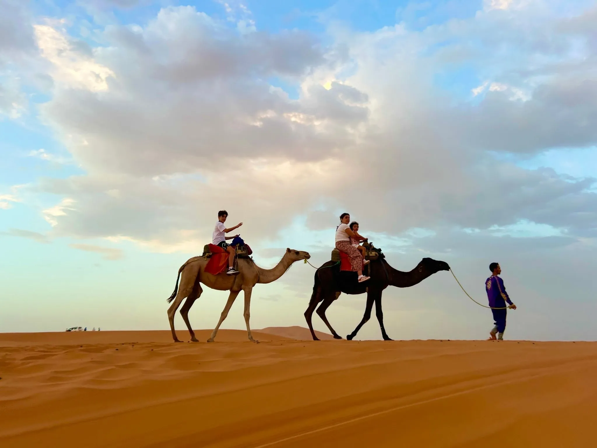 Family camel ride on Sahara dunes. A guide leads camels carrying riders across soft, orange sand beneath pastel clouds—and safe way to experience Morocco’s historical sites and natural beauty.