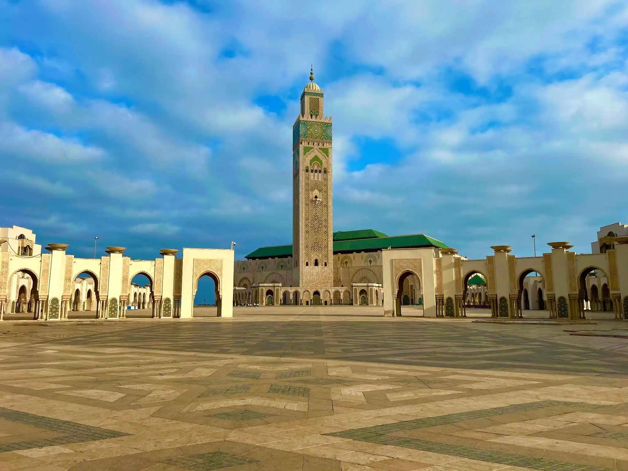 Hassan II Mosque (Casablanca). Wide esplanade and soaring minaret framed by arcades under a blue, cloud-dotted sky—making it one of the safest places to visit in Morocco.
