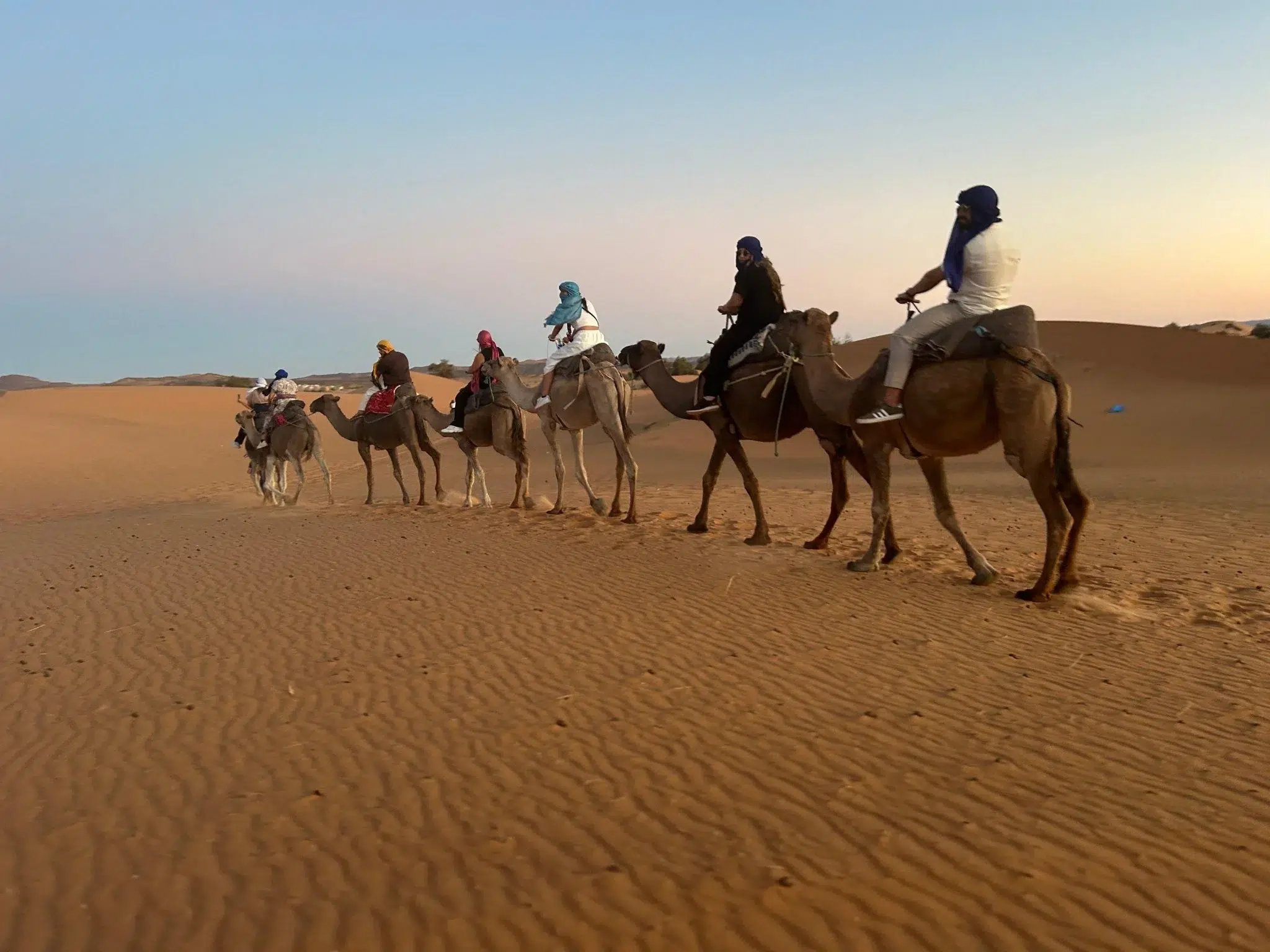 Line of travelers riding camels across the Erg Chebbi dunes at sunset during a 7-day Morocco desert and Chefchaouen tour.