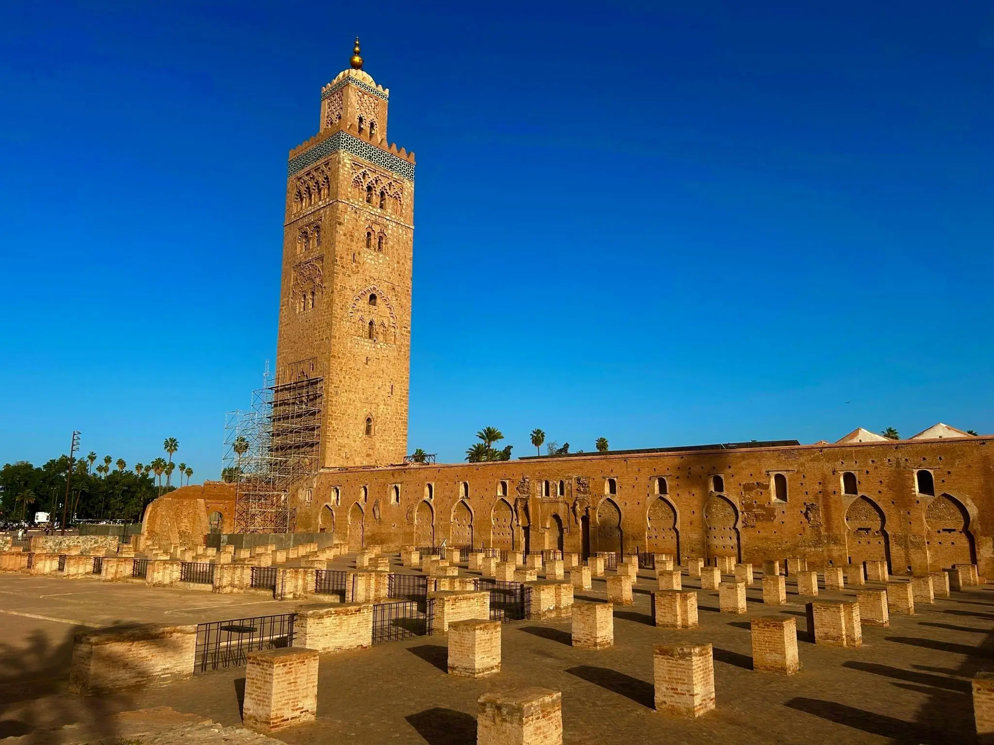 Wide view of the Koutoubia Mosque minaret and the ruins of the original mosque under a clear blue sky in Marrakech.